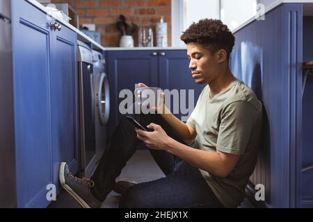 Seitenansicht eines jungen Birazialmannes, der zu Hause mit dem Smartphone im Küchengang einen Kaffeebecher hält Stockfoto