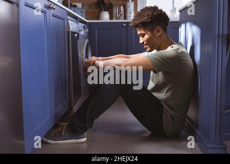 Volle Seitenansicht des jungen Birazialmannes, der zu Hause im Küchengang sitzt Stockfoto