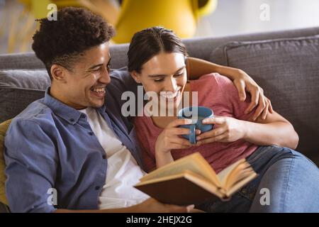 Glücklicher junger Biracial Mann liest Buch mit Freundin hält Kaffeebecher auf dem Sofa zu Hause Stockfoto