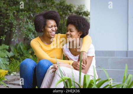 Glückliche Mutter, die mit Arm um die Teenager-Tochter im Hinterhof sitzt Stockfoto