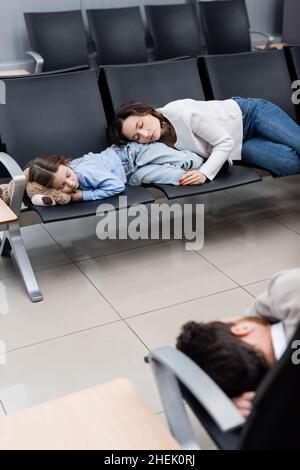 Mutter und Tochter schlafen auf den Sitzen am Flughafen in der Nähe eines verschwommenen Mannes in der Abflughalle