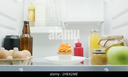 Leckerer Cupcake in der Nähe von Eiern, Flasche mit Getränken, Gläser mit Honig und Fruchtpüree im Kühlschrank Stockfoto