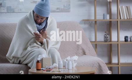 Kranker Mann, der auf dem Sofa in einer warmen Decke und einem Hut neben dem Tisch mit Papierservietten und Medikamenten sitzt Stockfoto