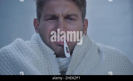 Unzufriedener kranker Mann mit Thermometer im Mund, der die Temperatur zu Hause misst Stockfoto