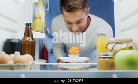 Lächelnder Mann, der Cupcake aus dem Kühlschrank in der Nähe von Getränken, Eiern, Gläsern und Behältern mit Lebensmitteln nahm Stockfoto