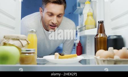 Ein zufriedener Mann, der sich leckeren Kuchen im Kühlschrank neben Getränken, Eiern und Saucen ansah Stockfoto