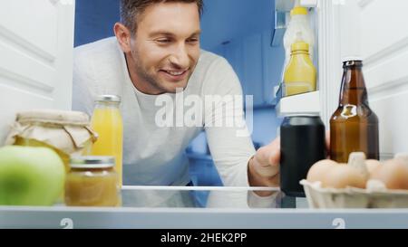 Glücklicher Mann, der Limonade aus dem Kühlschrank neben einer Flasche Bier, Orangensaft, Eiern und Gläsern mit Essen nahm Stockfoto