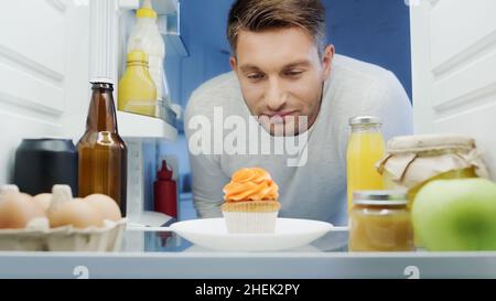 Mann, der köstlichen Cupcake in der Nähe von Getränken, Eiern und Saucen im Kühlschrank ansieht Stockfoto