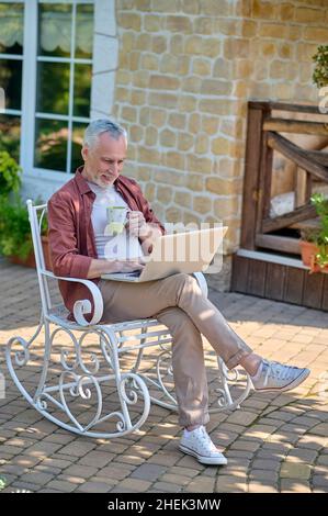 Grauhaariger Mann, der in einem Sessel sitzt und online etwas liest Stockfoto