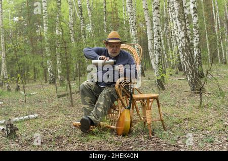 Bärtiger kaukasischer Senioren-Camper ruht sich im Birkenwald aus, sitzt auf einem Wicker-Schaukelstuhl und trinkt Tee aus der Thermosflasche Stockfoto