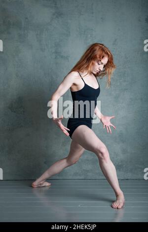 Petitie-Tänzerin mit fließendem rotbraunem Haar, die in schwarzen Shorts und Trikot im Fotostudio tanzt Stockfoto