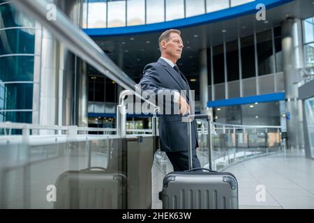 Ruhiger, nachdenklicher Geschäftsmann mit dem Koffer, der am Flughafenterminal steht Stockfoto