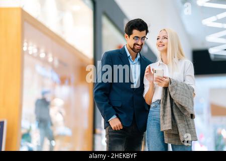 Fröhliches, schönes junges Paar, das in der Halle des Einkaufszentrums mit dem Mobiltelefon steht. Bärtiger Mann in Brille und blonde Frau beim Kauf im Ladengeschäft. Stockfoto