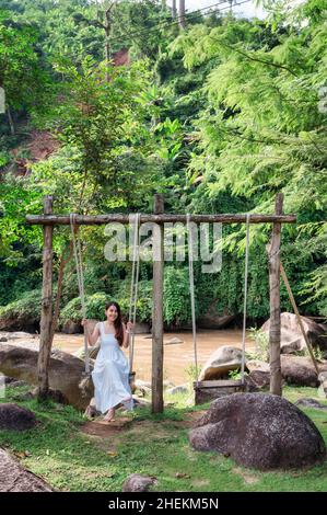 Glückliche asiatische Frau in weißem Kleid entspannende hölzerne Schaukel am Flussufer im Dschungel im Urlaub Stockfoto