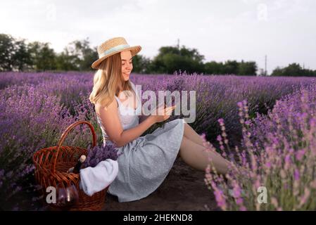 Ein junges schönes Mädchen, das zwischen den Lavendelbüschen sitzt und im Sommer bei Sonnenuntergang einen Blumenstrauß hält. Feld blühenden Lavendels und eine Dame Stockfoto