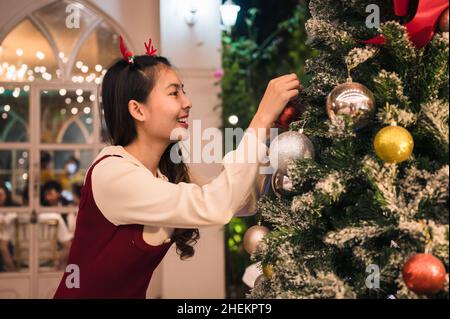 Fröhliche asiatische Frau trägt Geweih Stirnband Dekoration Ornament Ball auf weihnachtsbaum in der Nacht Stockfoto