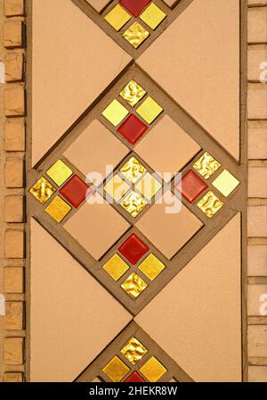 Fragment eines geometrischen, sich wiederholenden Musters aus einem Keramikmosaik an der Wand einer katholischen Kirche. Mosaikfliesen in Gold, Rot und Beige. Stockfoto