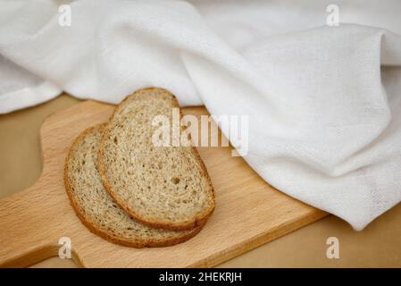 Zwei Scheiben Getreidebrot auf einem Holzbrett. Daneben befindet sich ein weißes Geschirrtuch.Beige Hintergrund, Draufsicht, Nahaufnahme. Gesundes Bio-Food-Konzept Stockfoto
