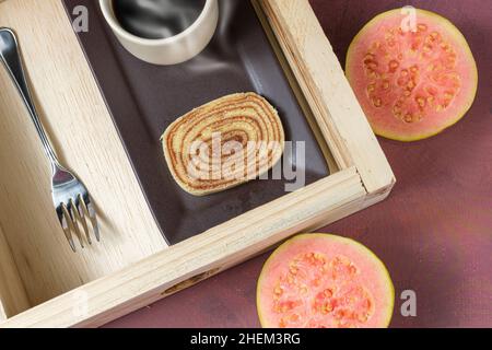 Scheibe Bolo de rolo auf einem braunen Teller, umgeben von Guava, einer Tasse Kaffee und einer Gabel. Stockfoto