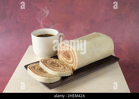 Geschnittenes Bolo de rolo auf einem braunen Teller, neben einer Tasse Kaffee. Stockfoto