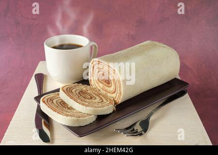 Bolo de rolo in Scheiben geschnitten auf einem braunen Teller, neben Besteck und einer Tasse Kaffee. Stockfoto