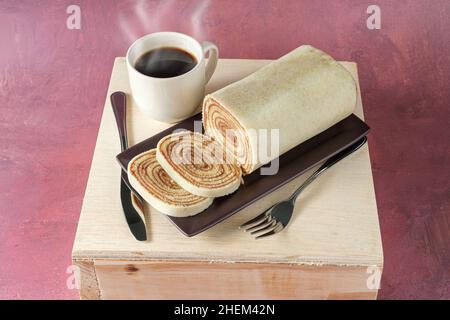 Bolo de rolo in Scheiben geschnitten auf einem braunen Teller, neben Besteck und einer Tasse Kaffee (Draufsicht). Stockfoto