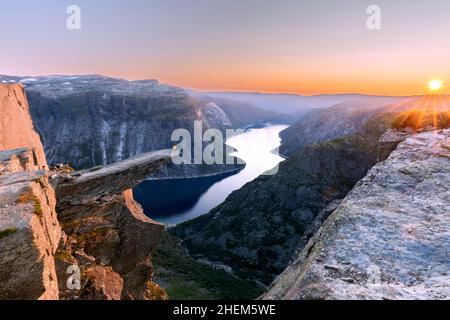 Ein Mann sitzt, während er seine Arme in die Luft wirft, am Felskante des Berges Trolltunga, der über Ringedalsvatnet thront und den Sonnenuntergang im Schnee beobachtet Stockfoto