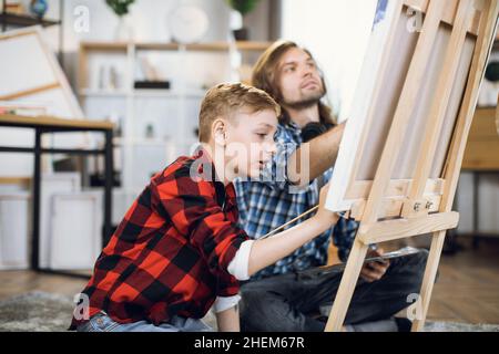 Kaukasischer Vater sitzt mit seinem Sohn auf dem Boden und lehrt, wie man mit Pinsel auf Staffelei malen. Kleiner Junge genießt Bildung Moment zu Hause. Entwicklungs- und Kreativitätskonzept. Stockfoto