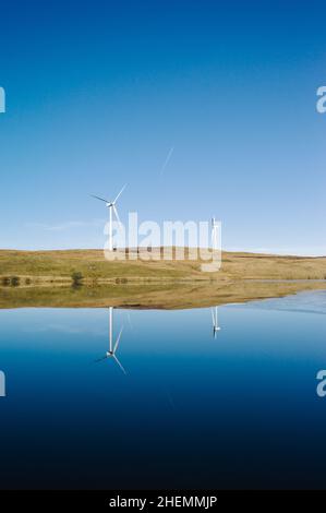 Zwei Windturbinen spiegeln sich an einem sonnigen Tag mit blauem Himmel in einem kleinen See oder loch wider. Stockfoto