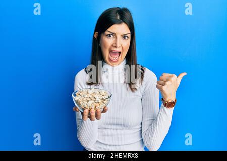 Junge hispanische Frau, die die Schale mit Kürbiskernen hält und den Daumen zur Seite zeigt, lächelt glücklich mit offenem Mund Stockfoto