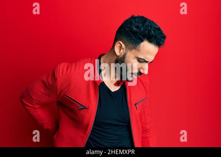 Junger hispanischer Mann mit roter Lederjacke, der Rückenschmerzen hatte, den Rücken mit der Hand berührte und muskuläre Schmerzen hatte Stockfoto