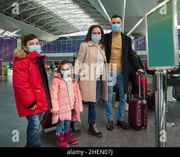 Glückliche kaukasische Familie von Vater, Mutter, Sohn und Tochter tragen schützende medizinische Maske in warmen Kleidern mit Gepäck und Koffern stehen gekleidet Stockfoto