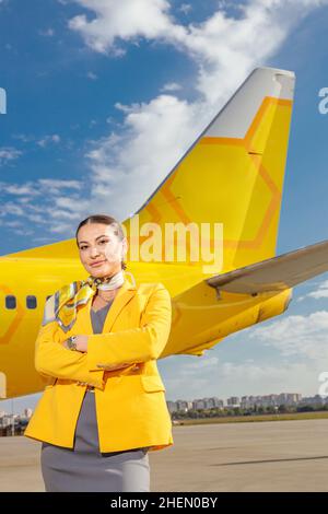 Fröhliche Flugbegleiterin in Stewardess-Uniform, die die Arme gekreuzt und lächelnd in der Nähe des Flugzeugs im Freien stehend hält Stockfoto