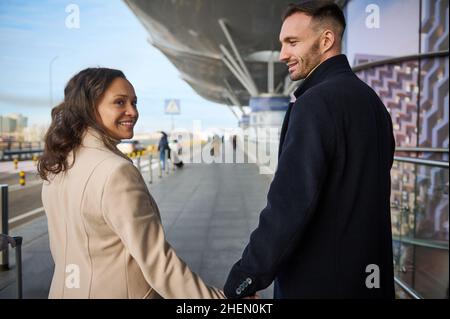 Liebevolles junges wiedervereinigtes multirassisches Paar, das sich anschaut, lächelt, die Hände hält, während es auf der Straße des internationalen Flughafens in Th geht Stockfoto