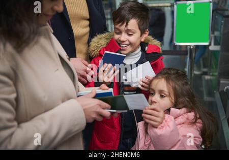 Fröhliche Kinder, die gerne mit der Familie reisen und ihrer Mutter Pässe geben, während sie am Flughafen Pass- und Zollkontrollen passieren. Flugreisen Stockfoto