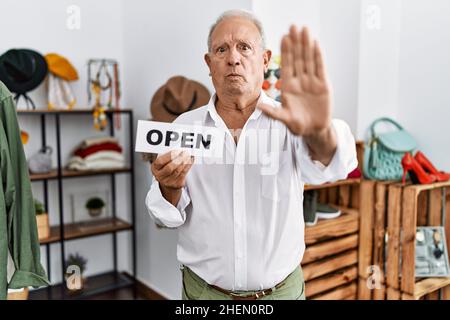 Ein älterer Mann hält ein Banner mit offenem Text im Einzelhandelsgeschäft mit offener Hand und tut ein Stoppschild mit ernsthaftem und selbstbewusster Äußerung, Verteidigungsgeste Stockfoto
