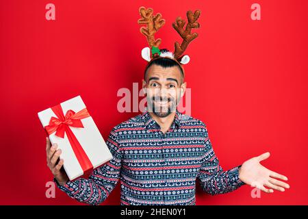 Junger hispanischer Mann trägt Hirsch weihnachtshut mit Geschenk feiert Leistung mit glücklichem Lächeln und Gewinner Ausdruck mit erhobener Hand Stockfoto