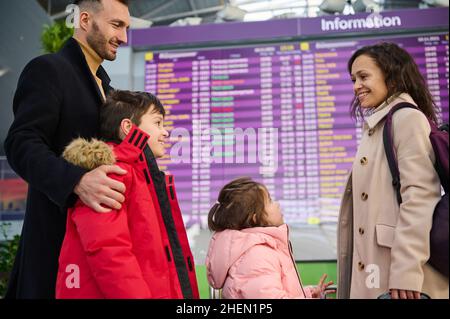 Eine glückliche, multirassische Familie, die gemeinsam vor dem Check-in-Board in der Abflughalle des Flughafens steht. Schöne Frau und Mann mit ihren Stockfoto
