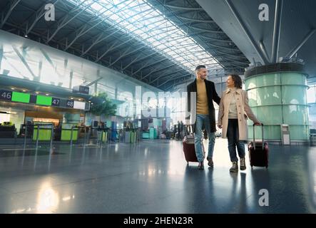 Porträt eines liebevollen Paares, das zusammen reist, Mann und Frau mit Koffern, die entlang der Abflughalle des internationalen Flughafens laufen Stockfoto