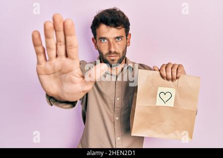 Schöner Mann mit Bart halten Lieferung Papiertasche mit Herz Erinnerung tun aufhören zu singen mit Handfläche der Hand. Warnung Ausdruck mit Negativ und ser Stockfoto