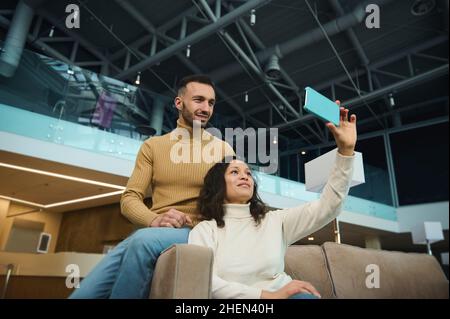 Charmante Frau und gutaussehender Mann, Freunde, Brautpaar, multirassische heterosexuelle Paare in der Liebe Selfie, während sie auf einen Flug in einem Inter zu gehen warten Stockfoto
