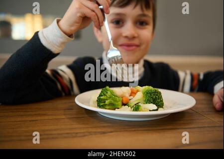 Das Bild konzentrierte sich auf einen Teller gedünstetes Gemüse. Entzückend lächelnder kaukasischer Teenager, der am Tisch sitzt und ein gesundes Mittagessen mit gedämpfter Karotte isst Stockfoto