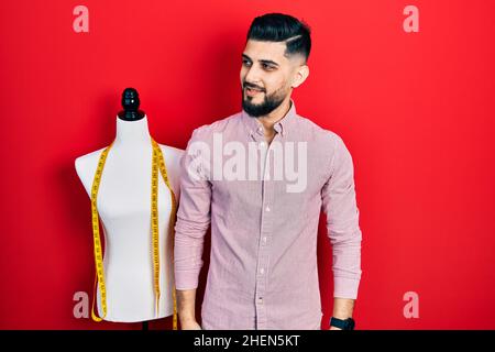 Hübscher Schneider mit Bart, der neben dem Übungspuppe steht und zur Seite schaut, entspannt die Profilpose mit natürlichem Gesicht und selbstbewusem Lächeln. Stockfoto