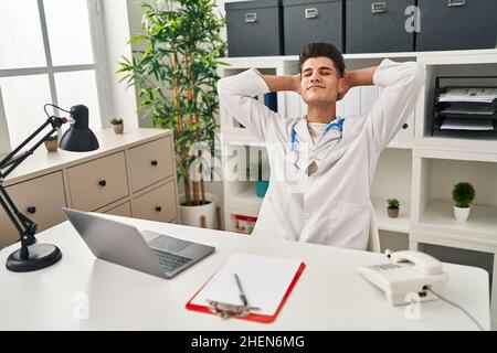 Der junge hispanische Mann in der Arztuniform entspannte sich in der Klinik mit den Händen am Kopf Stockfoto