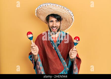 Junger hispanischer Mann mit mexikanischem Hut, der Maracas hielt, lächelte und lachte laut, weil er witzig verrückt scherzt. Stockfoto