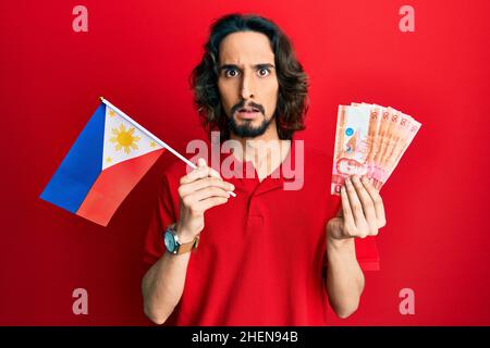 Junger hispanischer Mann mit philippinischer Flagge und Pesos-Banknoten, ahnungsloser und verwirrter Ausdruck. Zweifelskonzept. Stockfoto