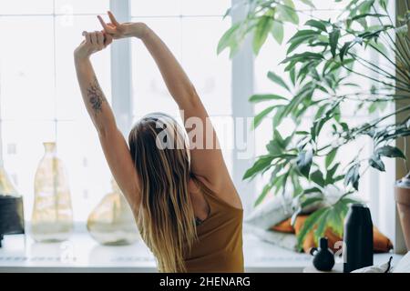 Frau mit blonden langen Haaren, die ein leuchtend gelbes Sporttop trägt, sitzt auf dem Bett mit weichen Kissen und blickt aus dem Fenster Stockfoto