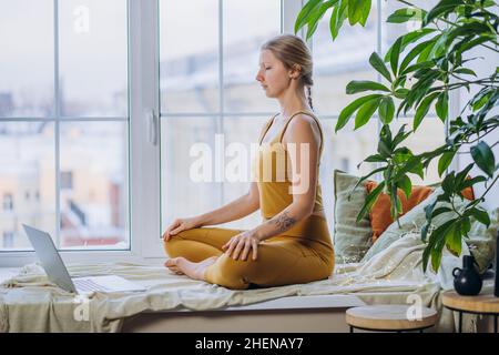 Die blonde junge Frau in gelbem Oberteil und Leggings meditiert in Yoga-Pose und sitzt auf einem mit Decke und Kissen bedeckten Fensterbrett mit einem Laptop Stockfoto