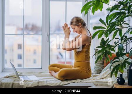 Die junge blonde langhaarige Frau in gelber Farbe meditiert in Lotuspose und sitzt auf einer Decke, die das Fensterbrett bedeckt, und schaut auf den Laptop-Bildschirm Stockfoto