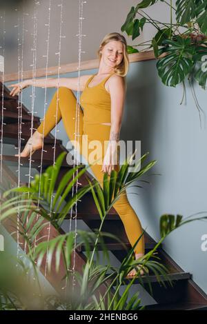 Blonde Frau mit gelbem Oberteil und Leggings macht vertikale Spalten, die sich auf einer Holztreppe in der Nähe von Girlande und grünen Pflanzen aufwärmen Stockfoto